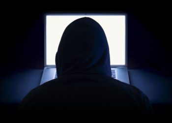 A hacker wearing a hoodie using a laptop in the dark.