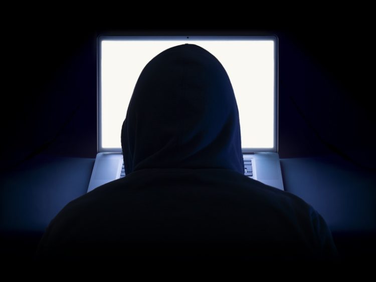 A hacker wearing a hoodie using a laptop in the dark.