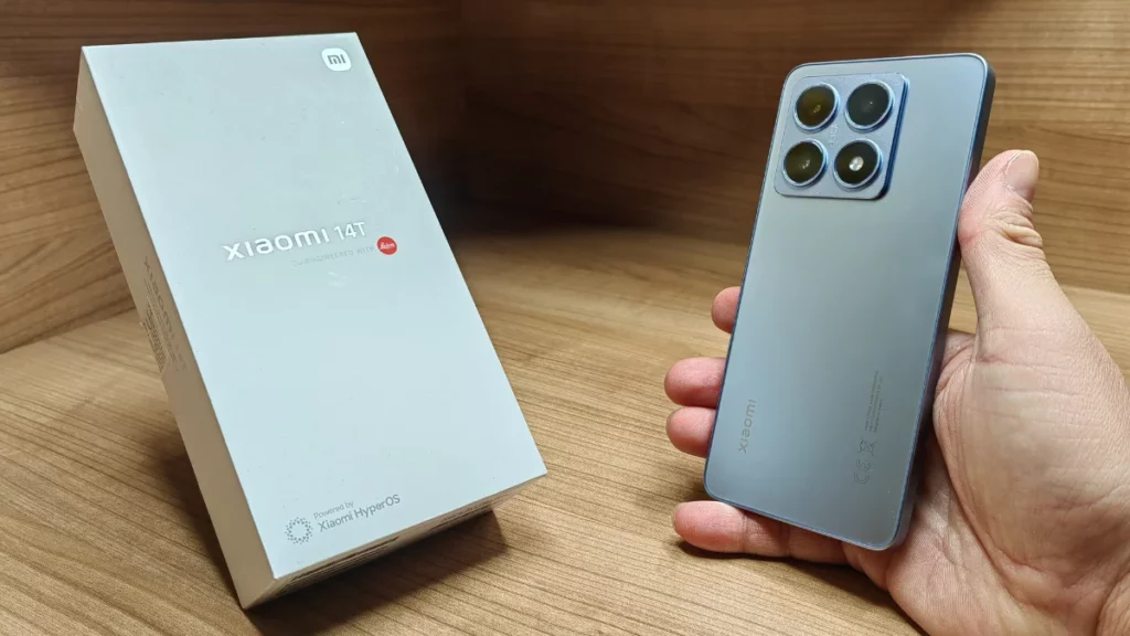 Tangan seseorang memegang HP Xiaomi 14T berwarna biru keabuan di samping kotak penjualan putihnya yang bertuliskan "CO-ENGINEERED WITH Leica".