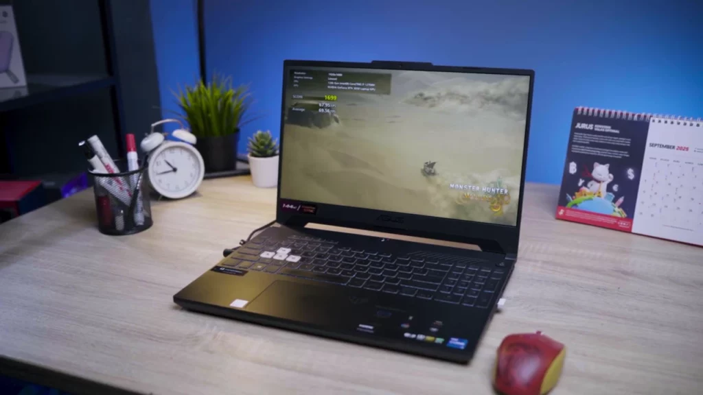 Tampilan laptop ASUS TUF F15 di atas meja kerja, menunjukkan desain sasis yang kokoh dan keyboard nyaman dengan numeric pad.