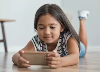 Seorang anak perempuan berambut panjang sedang berbaring tengkurap di lantai kayu, fokus menatap dan memegang ponsel pintar berwarna krem dengan kedua tangannya