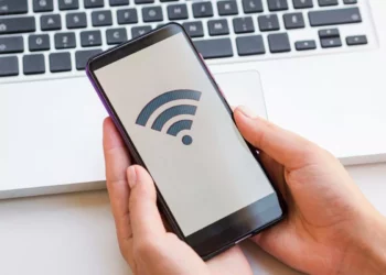 Tangan memegang smartphone yang menampilkan ikon sinyal WiFi di layar, dengan keyboard laptop di latar belakang.
