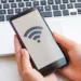 Tangan memegang smartphone yang menampilkan ikon sinyal WiFi di layar, dengan keyboard laptop di latar belakang.