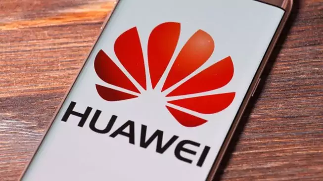 Layar smartphone menampilkan logo Huawei berwarna merah cerah, dengan latar belakang meja kayu gelap.