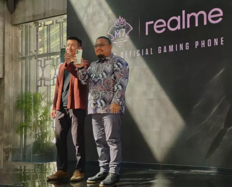 Dua pria, salah satunya Manajer Produk, berdiri di panggung memamerkan ponsel realme 15 Pro 5G, dengan latar belakang logo realme dan M7 Official Gaming Phone, dalam setting presentasi.