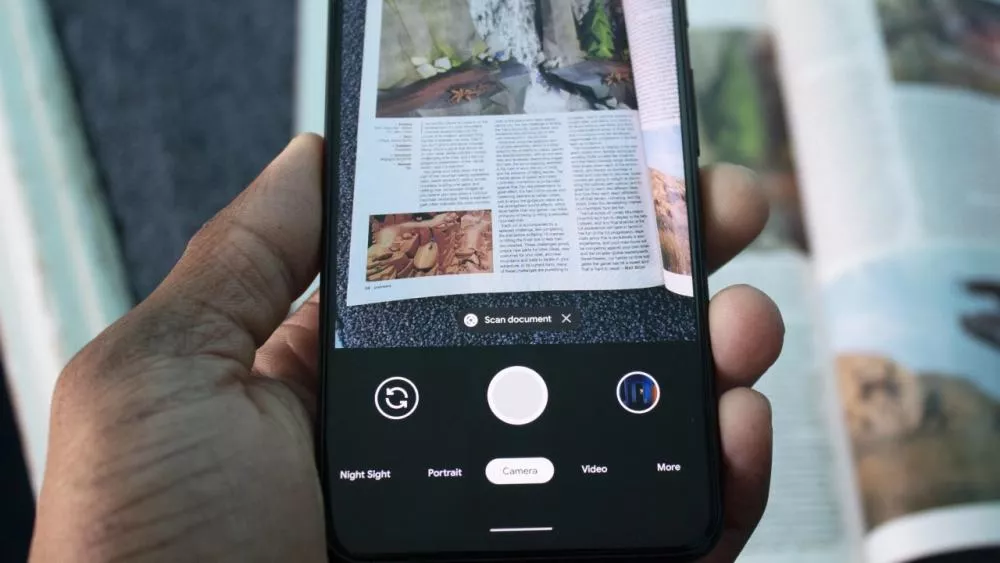 Tangan memegang smartphone yang menggunakan fitur kamera dengan mode "Scan document" yang diarahkan ke sebuah majalah atau buku.