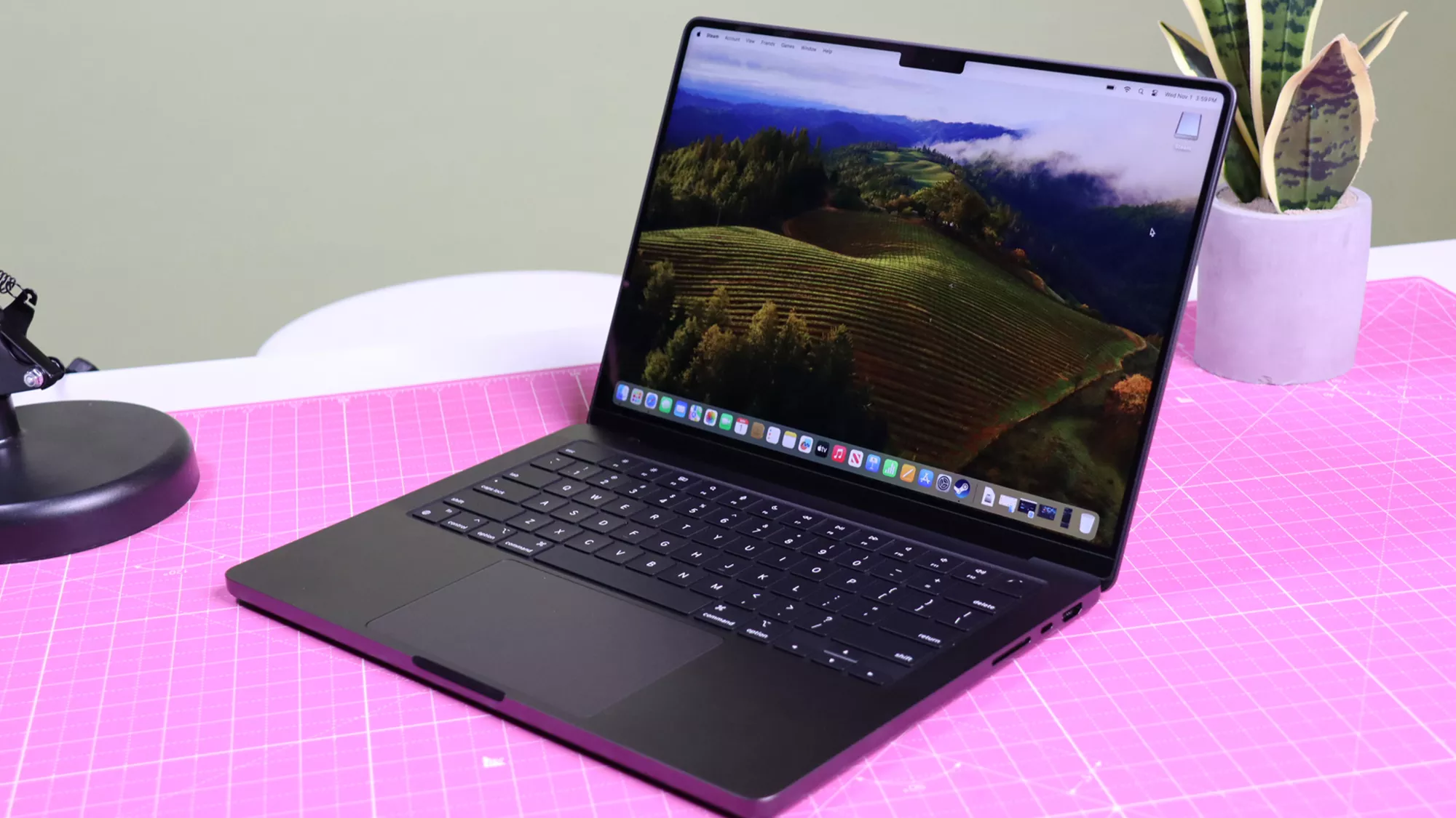 Laptop MacBook Pro M5 berwarna hitam diletakkan di atas meja pink, menampilkan layar beresolusi tinggi dengan pemandangan kebun anggur.