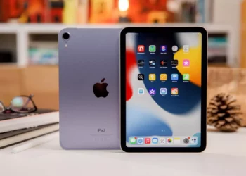 Perangkat iPad Mini berwarna ungu muda dengan logo Apple di bagian belakang, diletakkan di atas meja bersama dengan Apple Pencil dan kacamata.