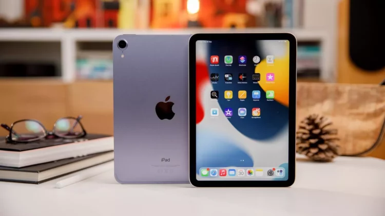 Perangkat iPad Mini berwarna ungu muda dengan logo Apple di bagian belakang, diletakkan di atas meja bersama dengan Apple Pencil dan kacamata.
