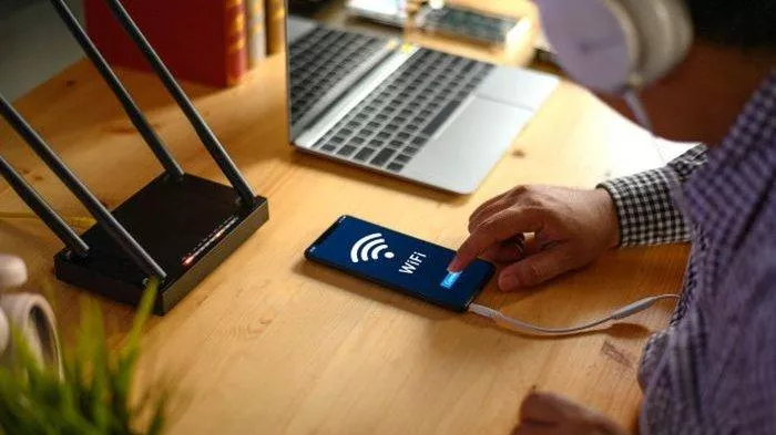 Tangan memegang smartphone yang menampilkan ikon sinyal WiFi di layar, dengan keyboard laptop di latar belakang.
