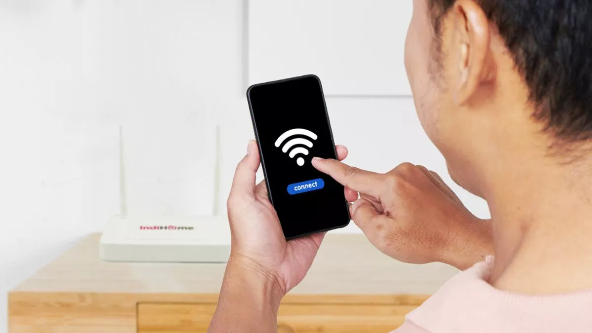 Seorang pria menyentuh layar smartphone untuk menyambungkan koneksi WiFi ke router IndiHome di latar belakang.
