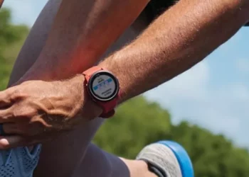 Lengan dan tangan seorang pelari memegang tali sepatu, dengan smartwatch berwarna merah yang menampilkan layar monitor detak jantung.