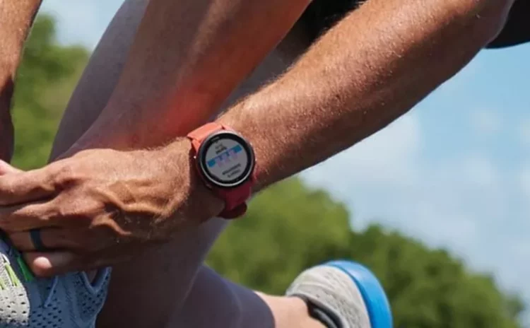 Lengan dan tangan seorang pelari memegang tali sepatu, dengan smartwatch berwarna merah yang menampilkan layar monitor detak jantung.