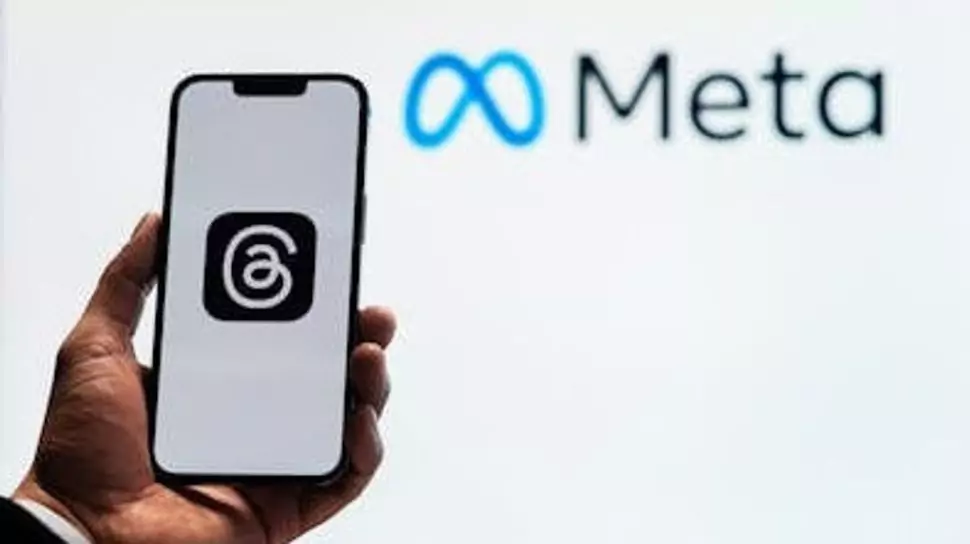 Tangan memegang smartphone yang menampilkan logo aplikasi Threads (@) di layar, dengan logo Meta di latar belakang putih.
