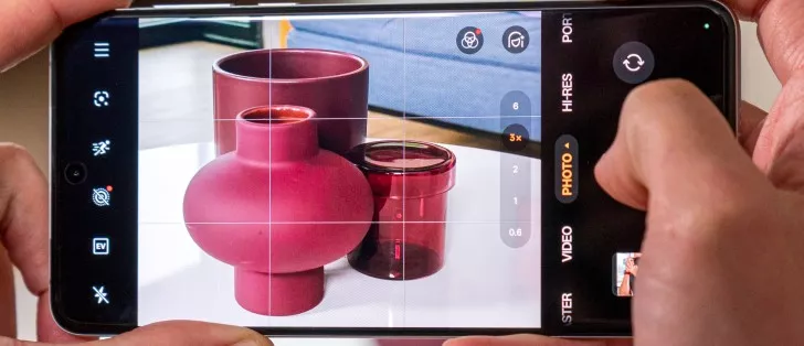 Tangan memegang smartphone yang menampilkan antarmuka kamera dengan objek vas merah dan opsi zoom 3x, menunjukkan fitur fotografi.