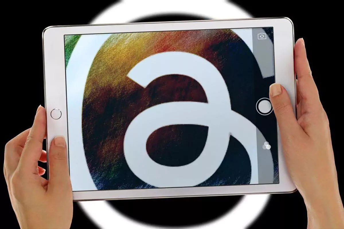 Sepasang tangan memegang tablet yang menampilkan logo aplikasi Threads (@) dengan efek visual warna-warni yang dinamis.