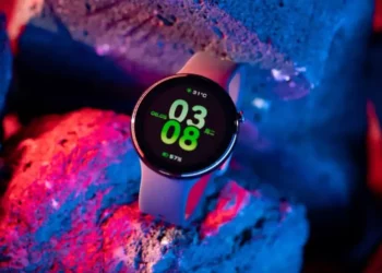Smartwatch Honor Watch Fit Active dengan tampilan antarmuka digital yang cerah, menunjukkan waktu, tanggal, dan suhu, diletakkan di antara bebatuan dengan pencahayaan neon biru dan merah