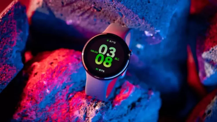 Smartwatch Honor Watch Fit Active dengan tampilan antarmuka digital yang cerah, menunjukkan waktu, tanggal, dan suhu, diletakkan di antara bebatuan dengan pencahayaan neon biru dan merah