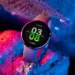 Smartwatch Honor Watch Fit Active dengan tampilan antarmuka digital yang cerah, menunjukkan waktu, tanggal, dan suhu, diletakkan di antara bebatuan dengan pencahayaan neon biru dan merah