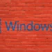 Logo dan tulisan "Windows 10" berwarna biru yang dicat di atas dinding bata merah, melambangkan sistem operasi yang sudah usang.