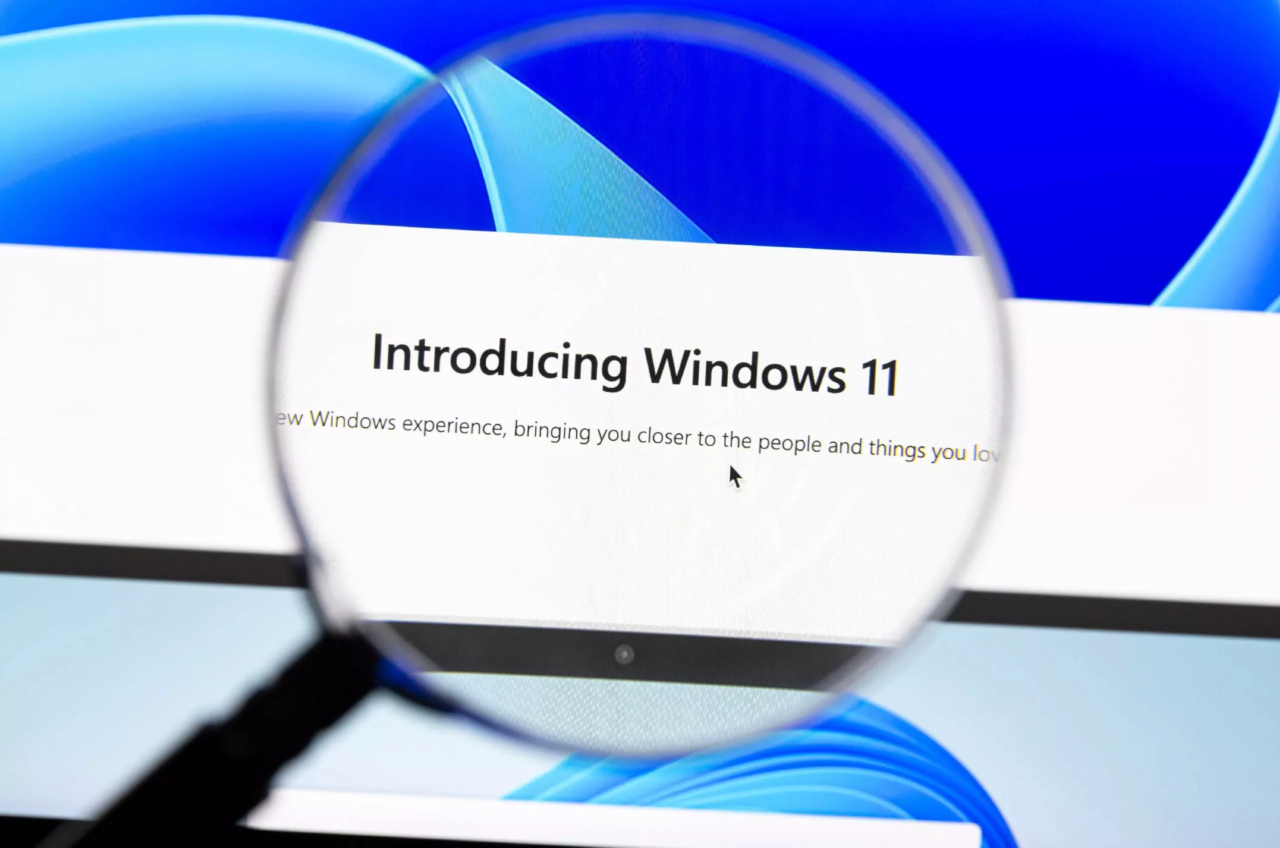 Layar komputer menunjukkan tulisan "Introducing Windows 11" melalui lensa pembesar, dengan wallpaper Windows 11 di latar belakang.
