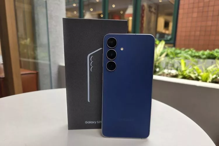 Tampilan belakang Samsung Galaxy S25 FE berwarna biru tua berdiri tegak di samping kotak kemasan berwarna hitam.
