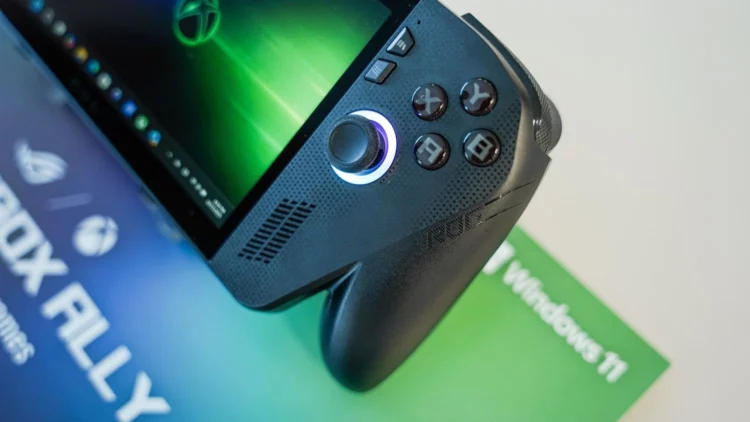 Sebuah foto close-up dari handheld gaming PC ROG Xbox Ally berwarna hitam, menyorot detail joystick kanan yang menyala dan tombol ABXY yang ikonik di atas materi promosi Windows 11.