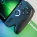 Sebuah foto close-up dari handheld gaming PC ROG Xbox Ally berwarna hitam, menyorot detail joystick kanan yang menyala dan tombol ABXY yang ikonik di atas materi promosi Windows 11.