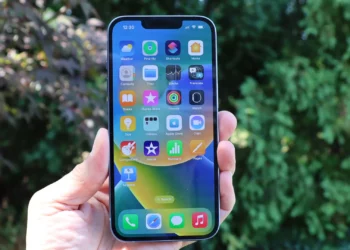 Tangan memegang iPhone dengan layar menyala menampilkan berbagai ikon aplikasi iOS di latar belakang hijau pepohonan, menyoroti antarmuka pengguna Apple yang familiar.