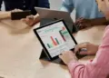 Seseorang mengetik di keyboard tablet yang menampilkan grafik data dan spreadsheet di layar, dalam suasana rapat.