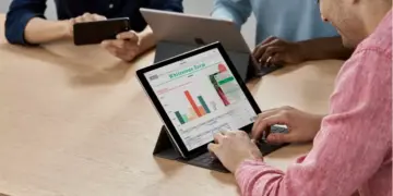 Seseorang mengetik di keyboard tablet yang menampilkan grafik data dan spreadsheet di layar, dalam suasana rapat.
