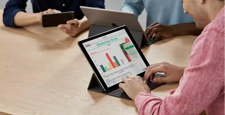 Seseorang mengetik di keyboard tablet yang menampilkan grafik data dan spreadsheet di layar, dalam suasana rapat.
