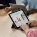Seseorang mengetik di keyboard tablet yang menampilkan grafik data dan spreadsheet di layar, dalam suasana rapat.
