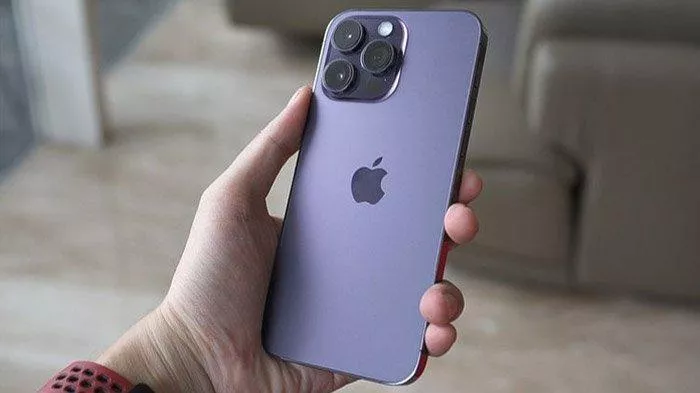 Tangan memegang bagian belakang iPhone berwarna ungu muda dengan tiga kamera menonjol, menampilkan logo Apple di tengah, menyoroti desain dan fitur kamera.
