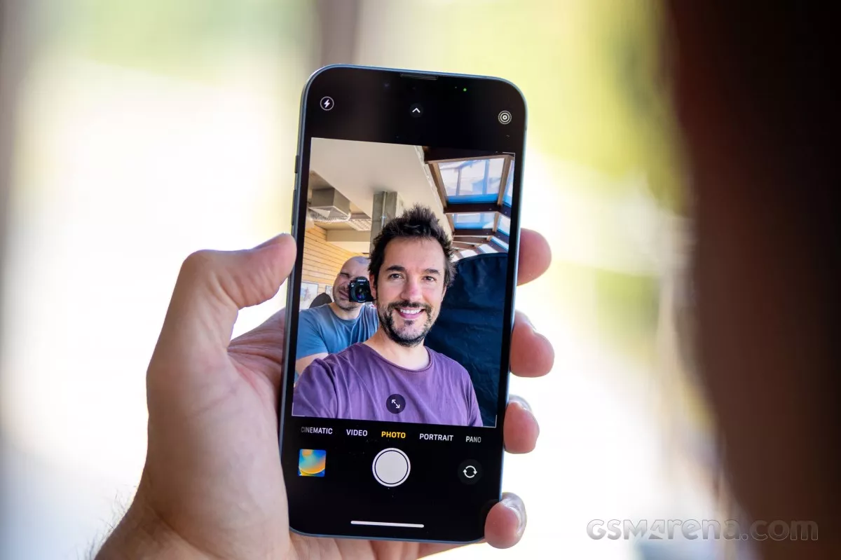 Tangan memegang iPhone yang menampilkan mode kamera selfie dengan seorang pria tersenyum di layar, menunjukkan fitur fotografi dan antarmuka kamera iOS.