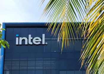 Logo Intel di fasad bangunan berwana biru gelap, dilihat dari balik daun pohon palem yang terang.