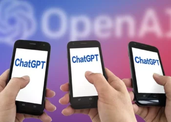 Tiga tangan memegang ponsel dengan tampilan aplikasi ChatGPT, di latar belakang buram logo OpenAI berwarna merah dan biru.