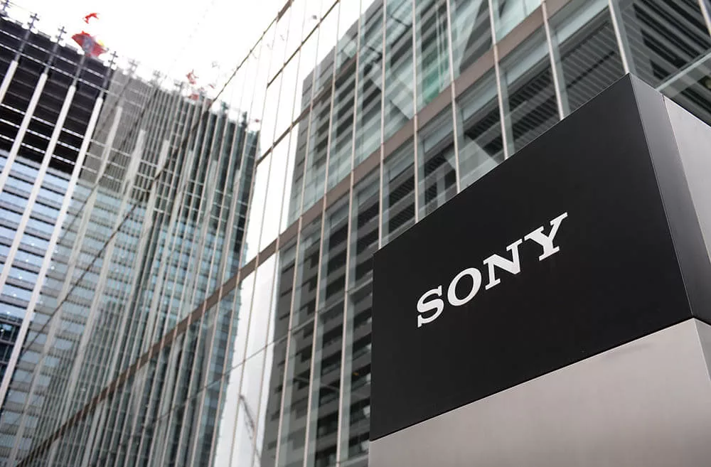 Logo perusahaan SONY berwarna putih pada papan nama hitam di luar gedung perkantoran modern yang terbuat dari kaca, menandakan lokasi perusahaan.