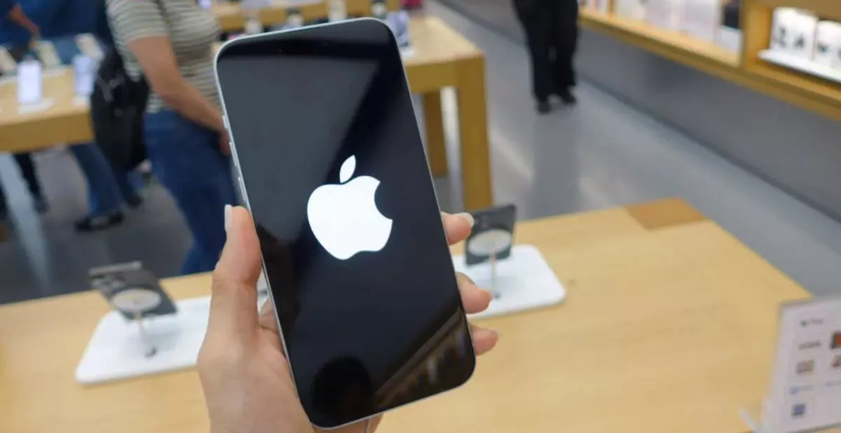 Tangan memegang iPhone baru yang sedang booting dan menampilkan logo Apple di tengah toko resmi Apple, mengacu pada peluncuran iPhone Edisi 20 Tahun.