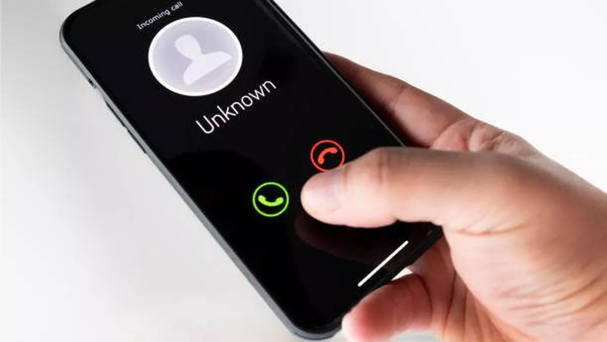 Tangan memegang smartphone yang menampilkan panggilan masuk dari "UNKNOWN", menunjukkan keraguan apakah harus menjawab atau tidak.