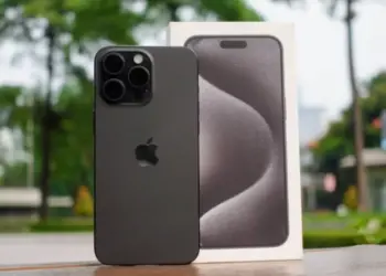Unit iPhone 15 Pro berwarna hitam matte yang diletakkan di samping kotak kemasan aslinya dengan latar belakang luar ruangan yang fokus.