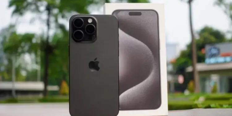 Unit iPhone 15 Pro berwarna hitam matte yang diletakkan di samping kotak kemasan aslinya dengan latar belakang luar ruangan yang fokus.