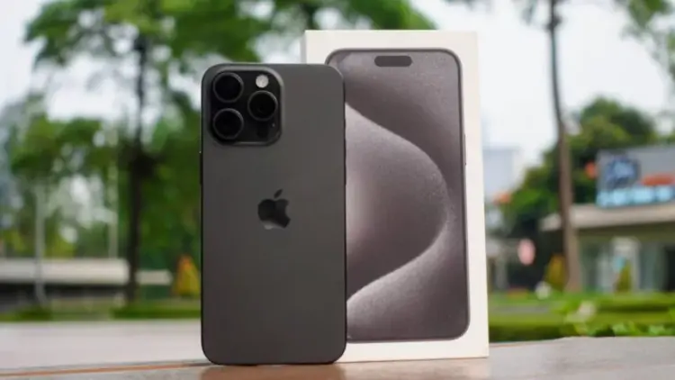 Unit iPhone 15 Pro berwarna hitam matte yang diletakkan di samping kotak kemasan aslinya dengan latar belakang luar ruangan yang fokus.