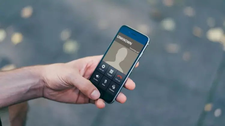 Close-up layar smartphone menunjukkan panggilan masuk dari "Unknown" dengan jari menekan tombol tolak (merah), mengisyaratkan bahaya.