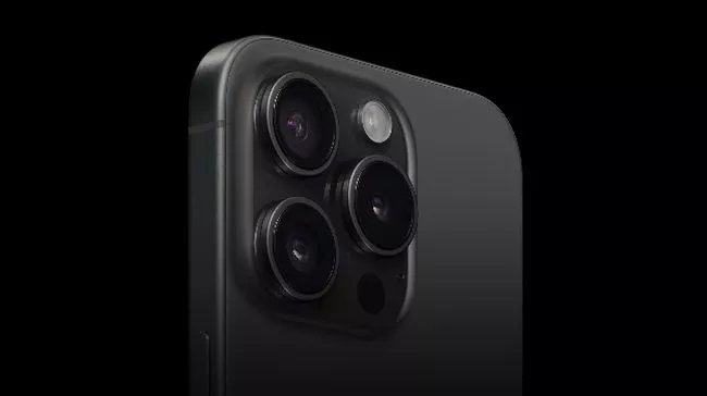 Close-up modul tiga kamera belakang iPhone 15 Pro berwarna hitam di latar belakang gelap, menonjolkan peningkatan spesifikasi.