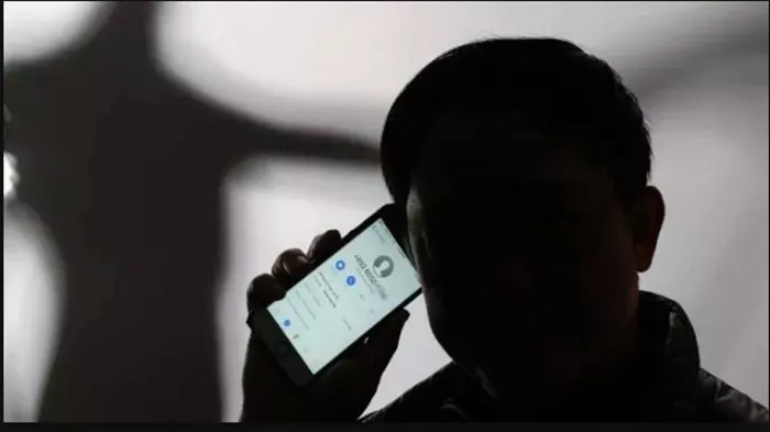 Siluet pria di ruangan gelap sedang berbicara di telepon dengan wajah tertutup bayangan, menyiratkan pelaku penipuan melalui panggilan misterius.