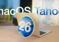 Logo macOS Tahoe dengan angka 26 di tengah diletakkan di depan laptop MacBook yang terbuka, menyoroti rilis update macOS Tahoe 26.2.
