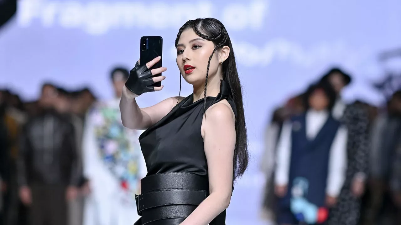 Model wanita berbusana hitam memegang smartphone di catwalk fashion show, menyoroti aspek gaya dan kemewahan.
