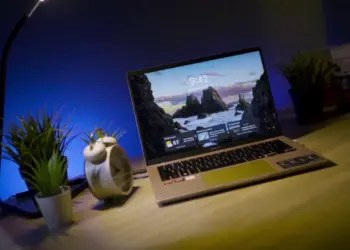 Laptop ASUS Vivobook 14 (M1407) menyala di atas meja kayu dengan latar belakang pencahayaan biru ambient dan tanaman. Gambar unggulan untuk artikel review laptop AI 10 jutaan yang memiliki daya tahan baterai 15 jam.