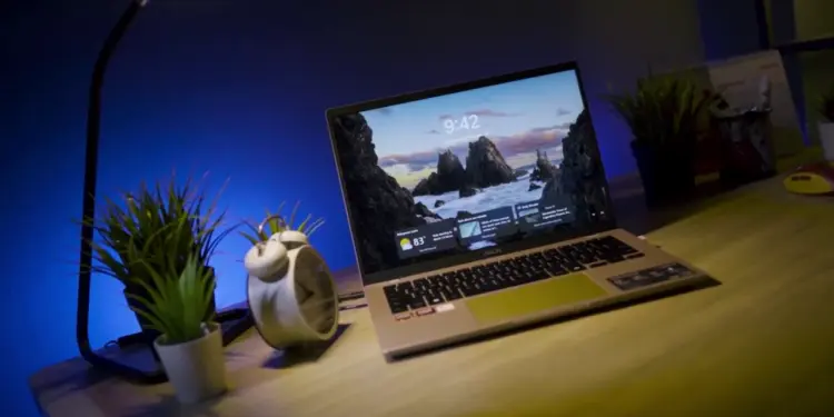 Laptop ASUS Vivobook 14 (M1407) menyala di atas meja kayu dengan latar belakang pencahayaan biru ambient dan tanaman. Gambar unggulan untuk artikel review laptop AI 10 jutaan yang memiliki daya tahan baterai 15 jam.